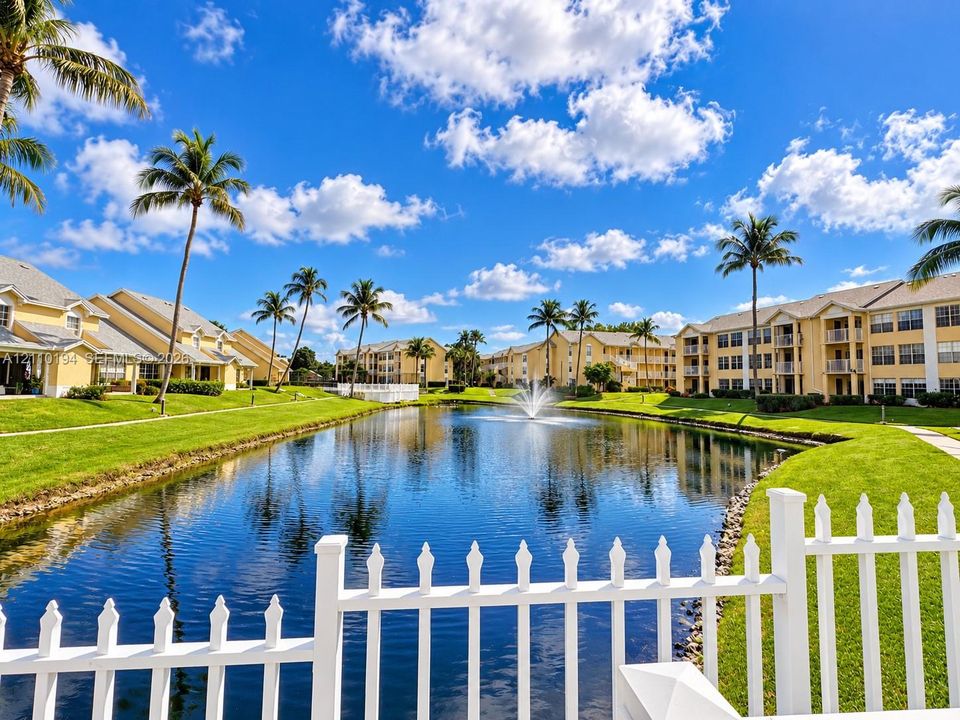 Community pond featuring a central fountain, manicured lawn, white picket fencing, and surrounding palm trees