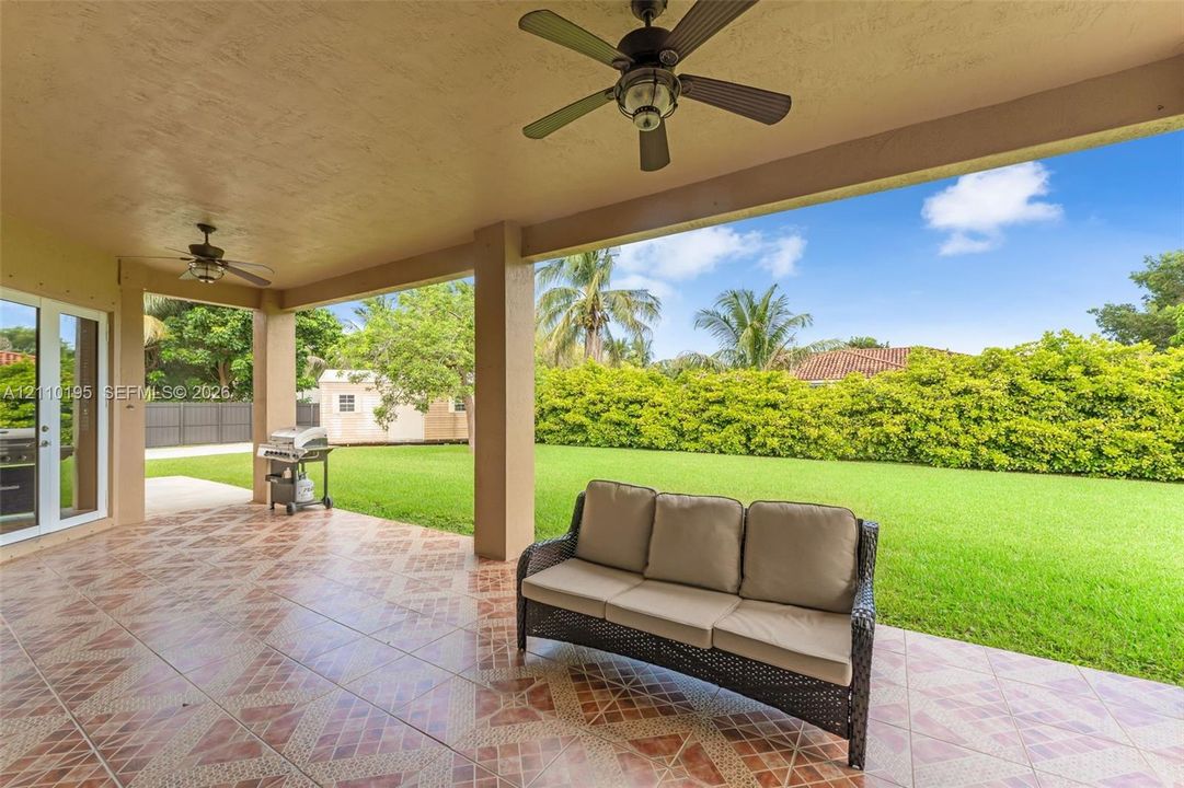 Covered patio featuring patterned tile flooring, two ceiling fans, and a spacious lawn area with established greenery