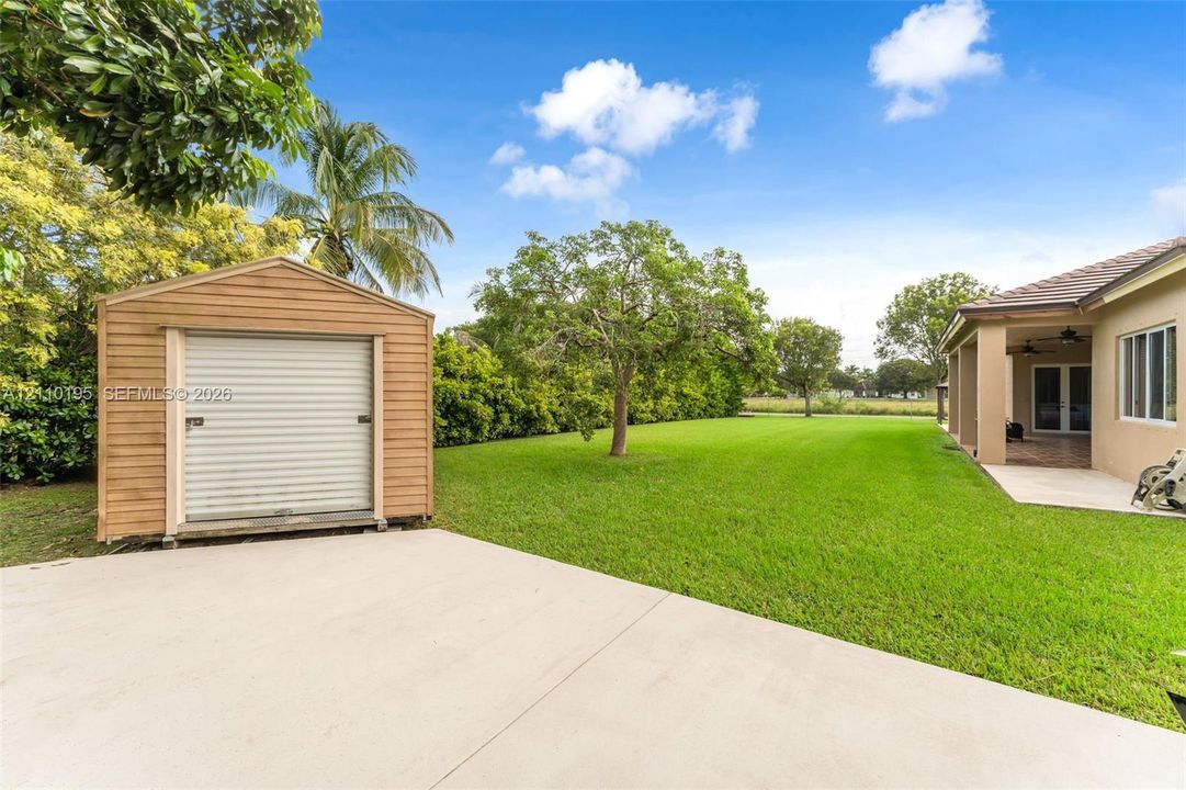 Expansive backyard featuring a concrete patio, detached wood-finish storage shed, mature trees, and manicured lawn