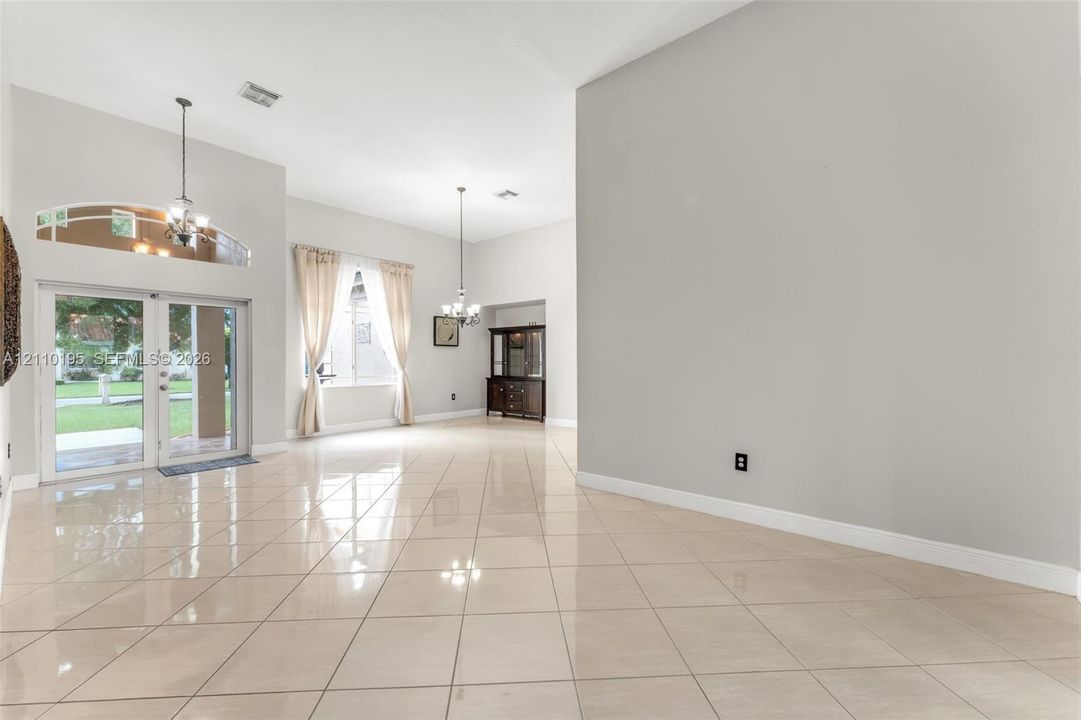 High-ceilinged interior featuring polished ceramic tile flooring, dual multi-light chandeliers, arched transom window, and double glass-paneled exterior doors