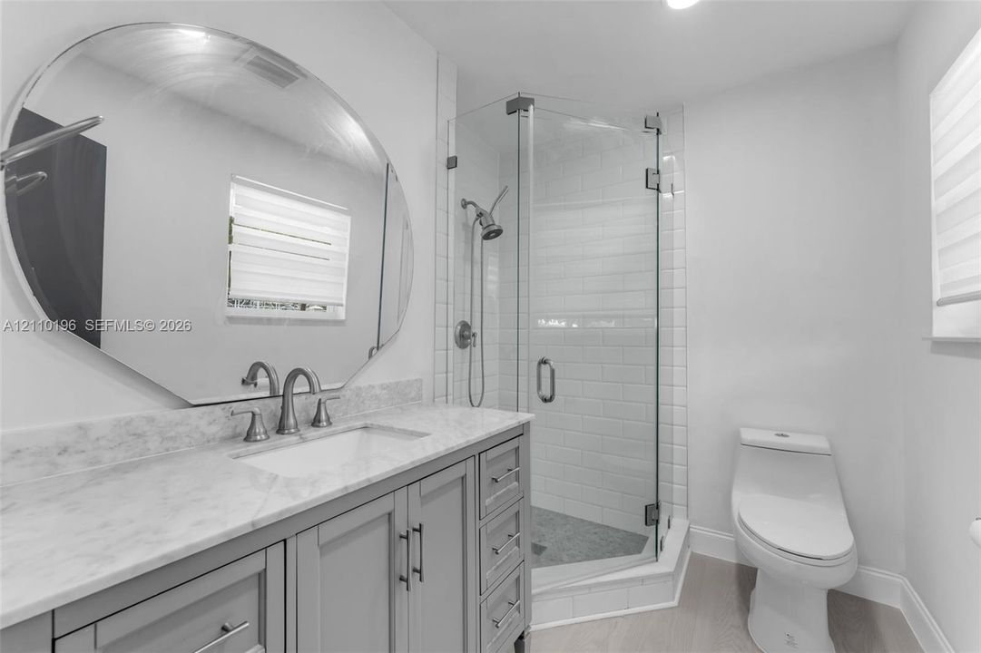 Bathroom featuring a gray vanity with a neutral stone countertop, an oversized circular mirror, a glass-enclosed shower with subway tile surround, and wood-finish flooring