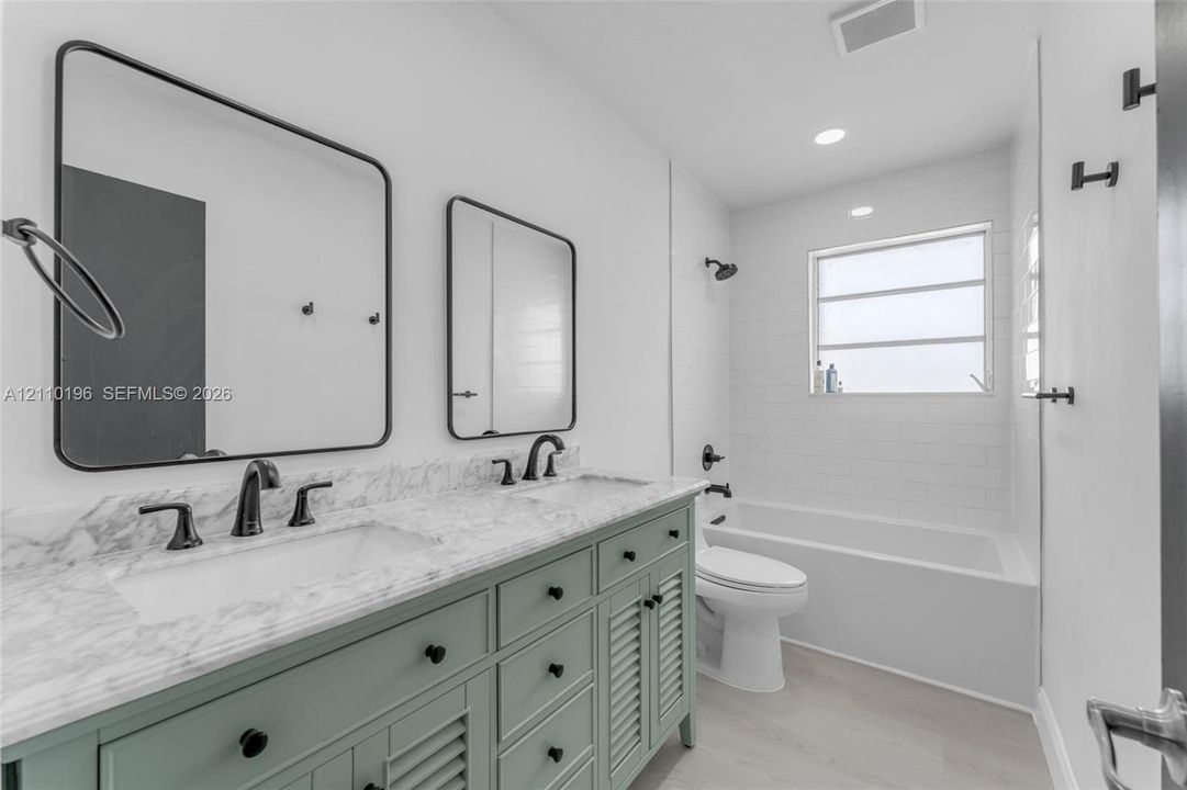 Double vanity with stone-look countertop, green cabinetry with louvered door detailing, matte black fixtures, rectangular wall-mounted mirrors, and a tub-shower combination with white subway tile surround