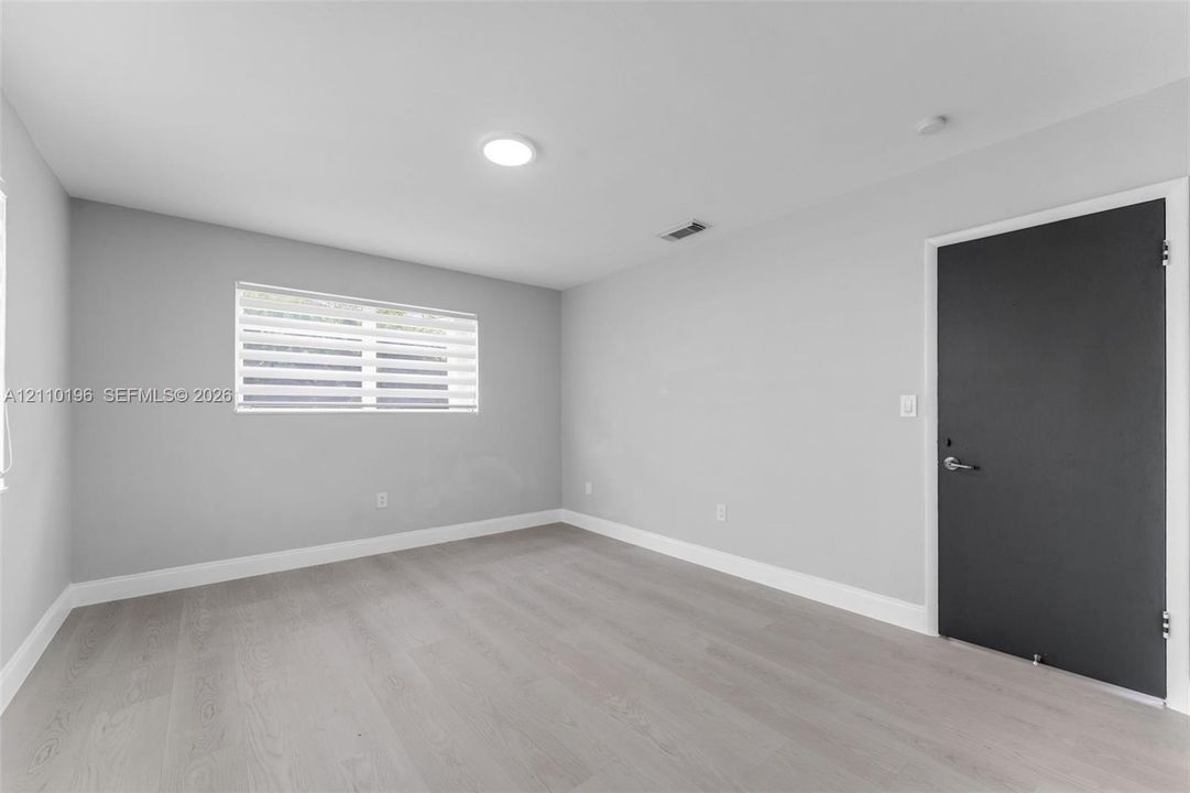 Neutral-toned interior space featuring wood-finish flooring, light gray walls, and a contrasting dark-finished door with metallic hardware