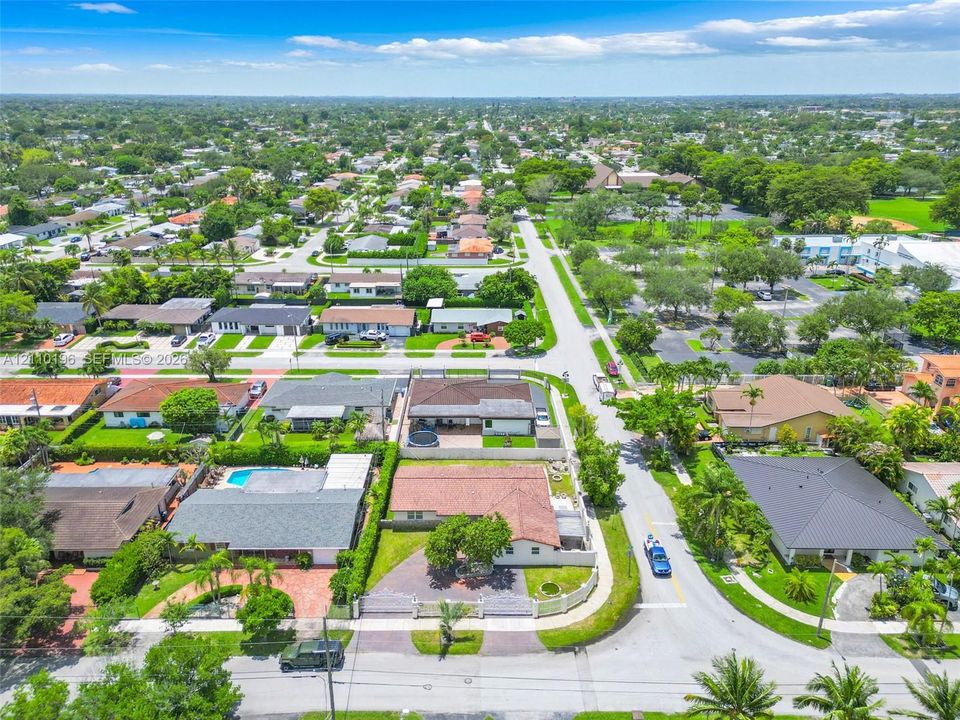 Aerial perspective of a corner lot property featuring a paved driveway, enclosed perimeter wall, mature landscaping, and a multi-section shingle roof