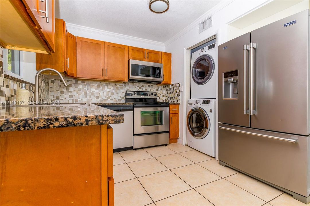 Kitchen featuring wood cabinetry, stone-finish countertops, mosaic tile backsplash, stainless steel appliances, and built-in stacked laundry