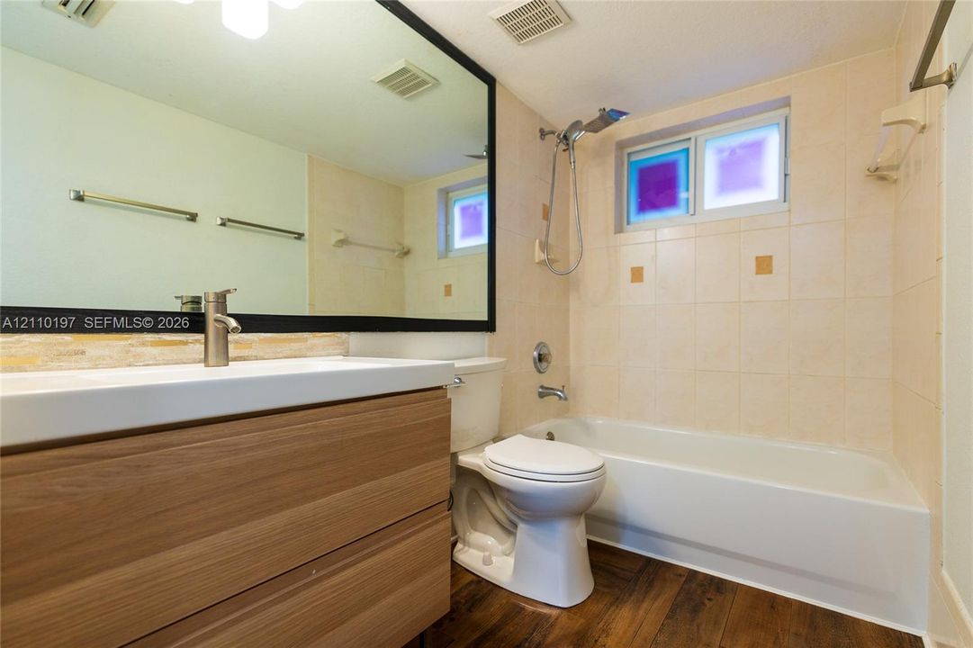 Bathroom featuring a wood-finish vanity with a rectangular sink, a large wall-mounted mirror with a black frame, a tub-shower combination with tile surround, and wood-finish flooring
