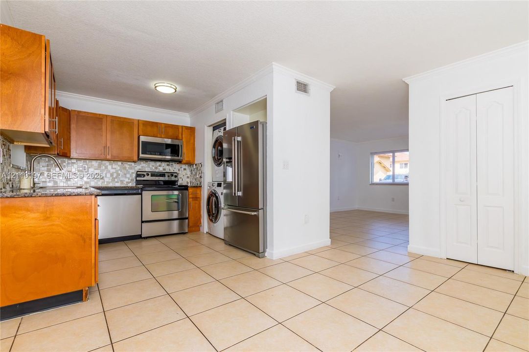 Kitchen featuring wood cabinetry, mosaic tile backsplash, stainless steel appliances, tile flooring, and a built-in laundry closet with stacked washer and dryer