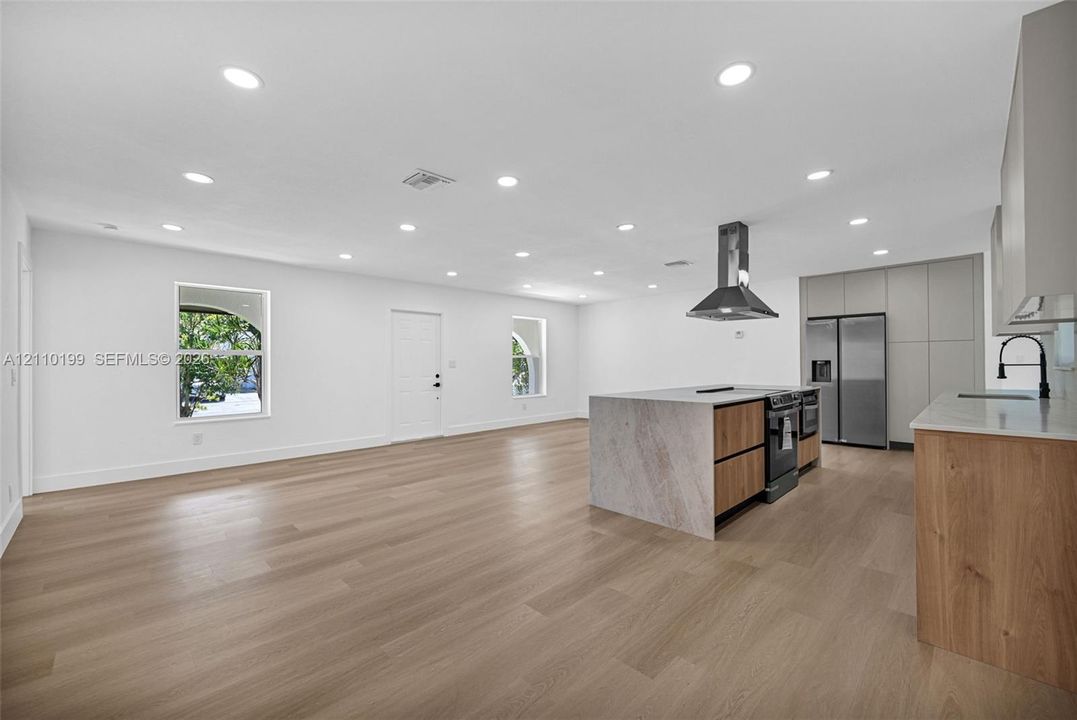 Open-concept interior featuring light wood-finish flooring, recessed lighting, a kitchen island with stone-finish waterfall counters, flat-panel wood cabinetry, and stainless steel appliances including a range hood