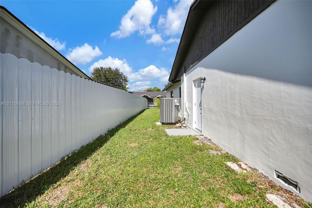 Side yard featuring a white corrugated metal fence, grass lawn, and exterior wall-mounted lighting