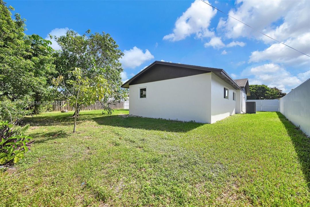 Exterior side profile featuring light-toned stucco siding, a low-pitched roof with dark-toned gable end, and a spacious grass yard enclosed by a corrugated metal fence