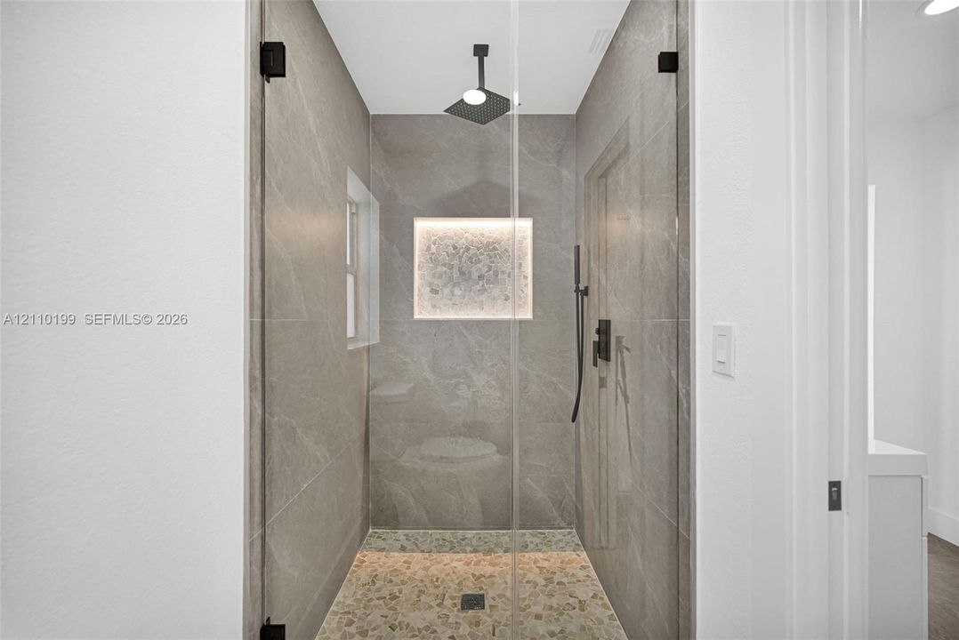 Walk-in shower featuring large-format tile surround, frameless glass enclosure, rainfall showerhead, pebble-finish flooring, and a built-in recessed niche with integrated lighting