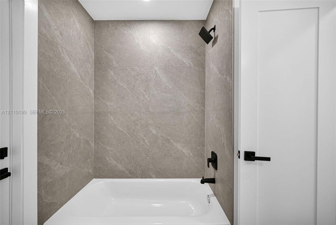 Full-height neutral stone-finish tile surround, white soaking tub, and matte black plumbing fixtures