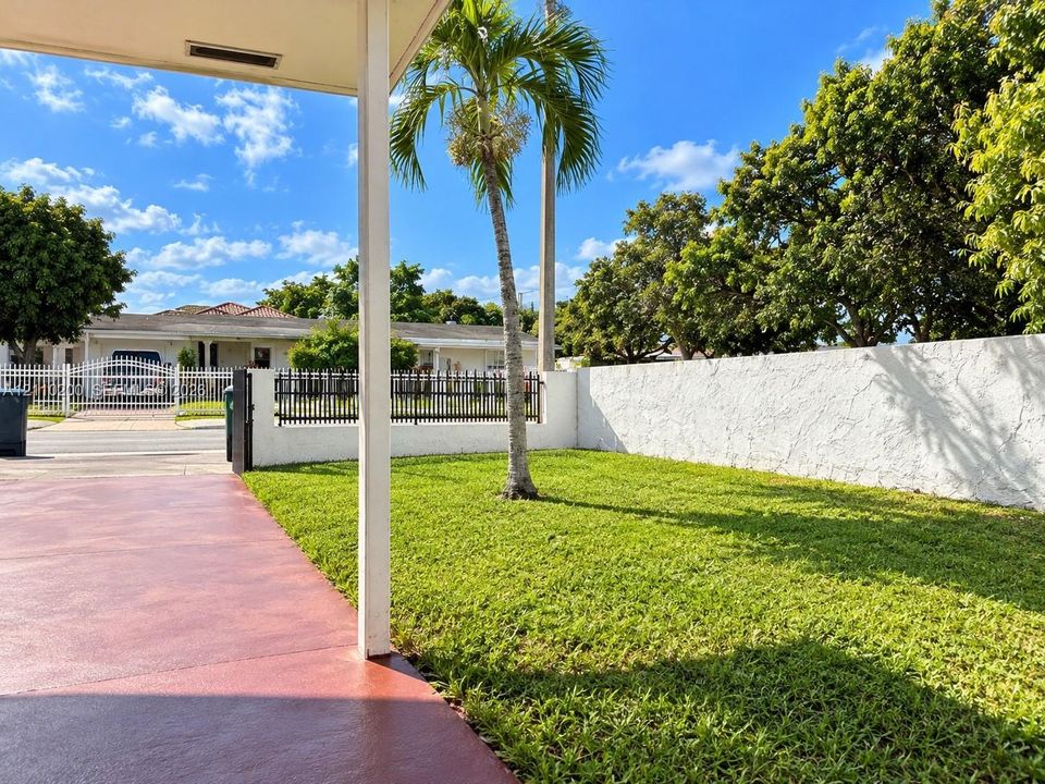 Covered patio with a concrete surface, manicured lawn, perimeter stucco wall, metal fencing, and mature palm tree