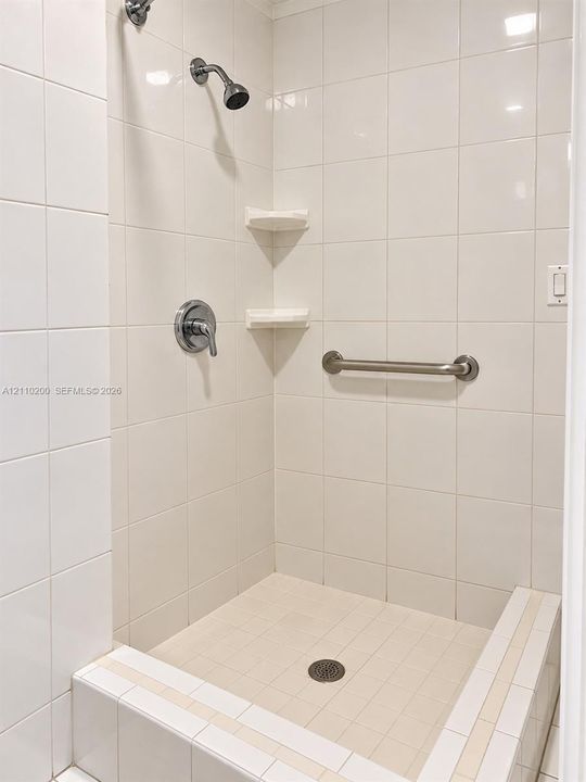 Shower stall featuring white as wall and floor tiling, chrome fixtures, integrated corner shelving, and a metal grab bar