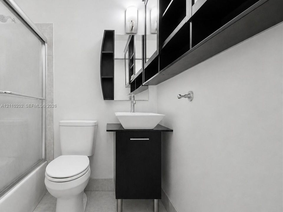 Modern vanity featuring a dark-finish base, square vessel sink, and chrome faucet, accompanied by built-in wall-mounted cabinetry and a glass-enclosed tub shower