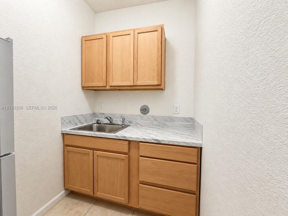 Wood-finish cabinetry featuring upper and lower storage, stone-patterned laminate countertop, stainless steel drop-in sink with chrome faucet, and neutral tile flooring