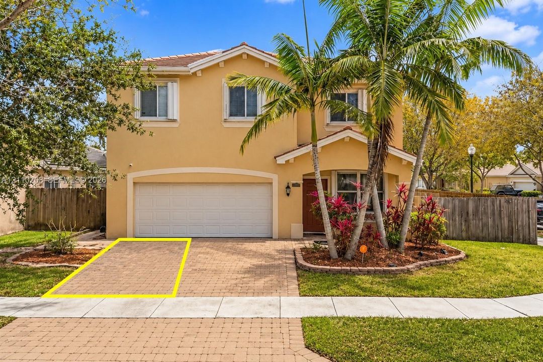 Two-story stucco exterior featuring a paved stone driveway, an integrated two-car garage, and landscaped garden beds with mature palms