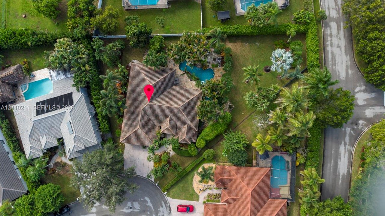Aerial view featuring a multi-hipped roofline, an in-ground swimming pool, mature palm trees, and landscaped grounds