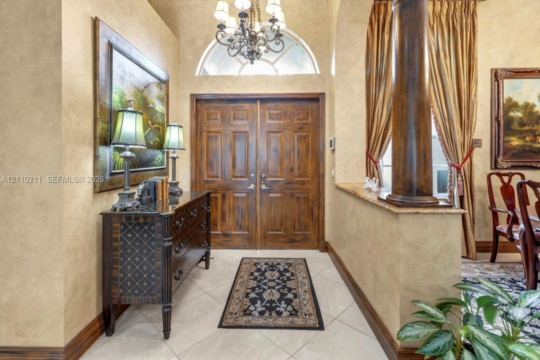 Foyer featuring double wood entry doors, an arched transom window, a decorative chandelier, tile flooring, and a stone-topped half wall with an ornamental column