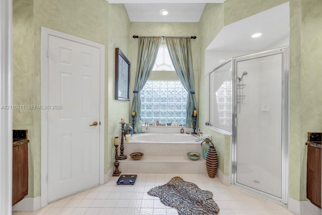Bathroom featuring a recessed soaking tub with a tiled surround, glass block window, separate shower stall with a glass enclosure, and dual cabinetry with dark stone countertops