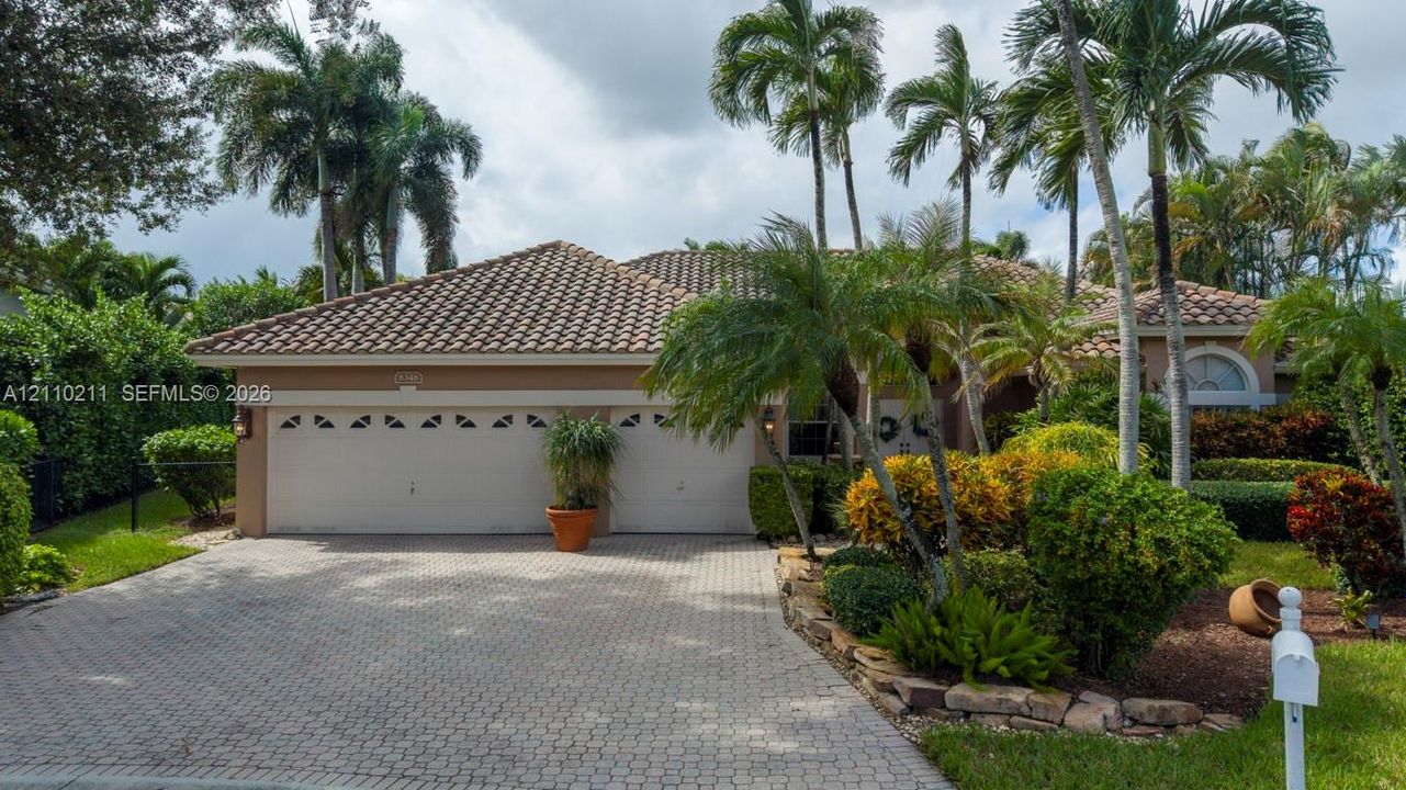 Single-story exterior featuring a clay tile roof, two-car garage with arched windows, and a paver stone driveway surrounded by mature palms and professional landscaping