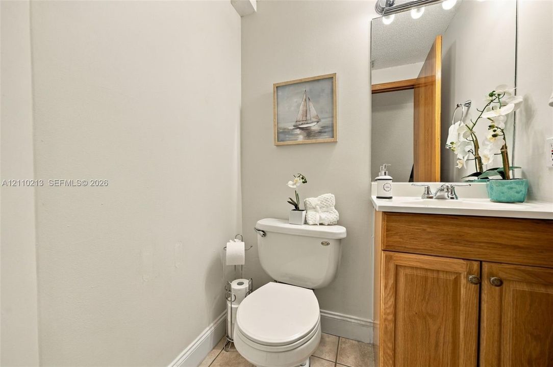 Wood-finish cabinetry, solid-surface countertop with integrated basin, polished chrome faucet, full-width vanity mirror, and ceramic tile flooring