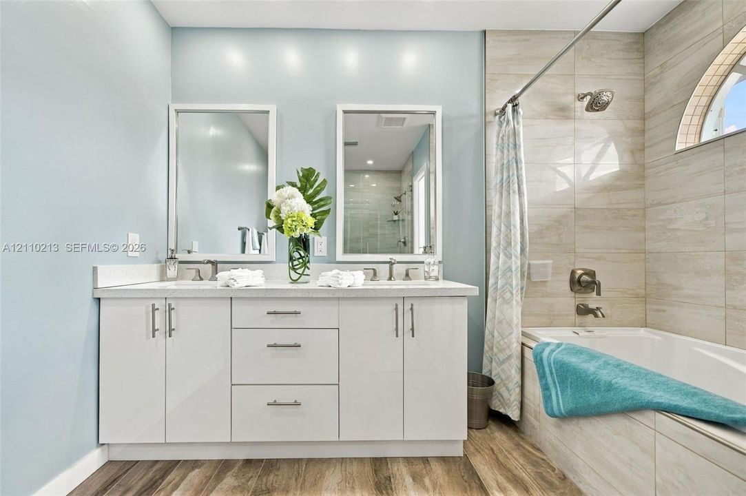 Dual-sink vanity with white cabinetry and light-toned solid surface countertop, wood-finish flooring, full-height tile shower surround, and semi-circular window