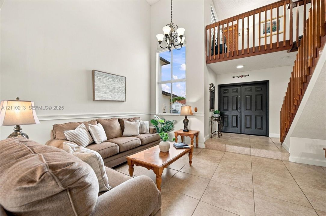 High-ceilinged interior featuring large-format tile flooring, a multi-pane window, a traditional hanging chandelier, and an elevated mezzanine with wooden railings