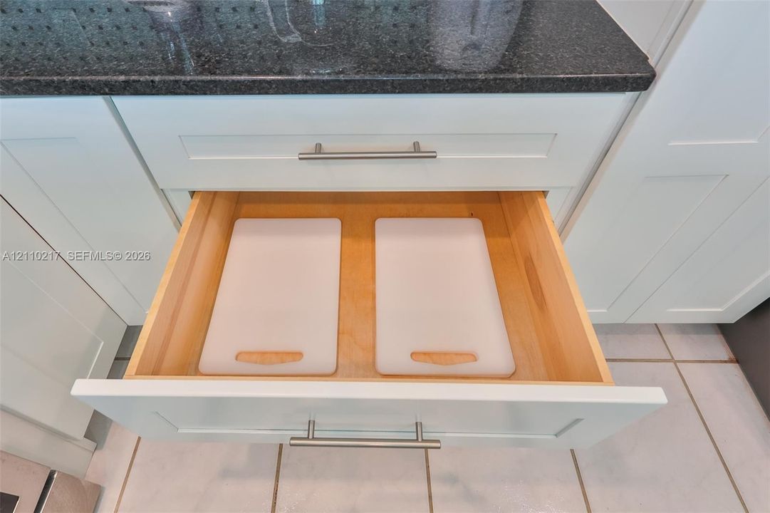 Shaker-style white cabinetry featuring a pull-out drawer with a light-finish wood interior, brushed-metal hardware, and dark stone-finish countertop
