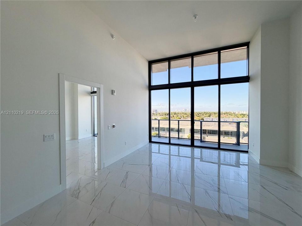 High-ceiling interior featuring polished stone-finish floor tiling, expansive floor-to-ceiling windows with black metal framing, and a balcony with a glass and metal railing