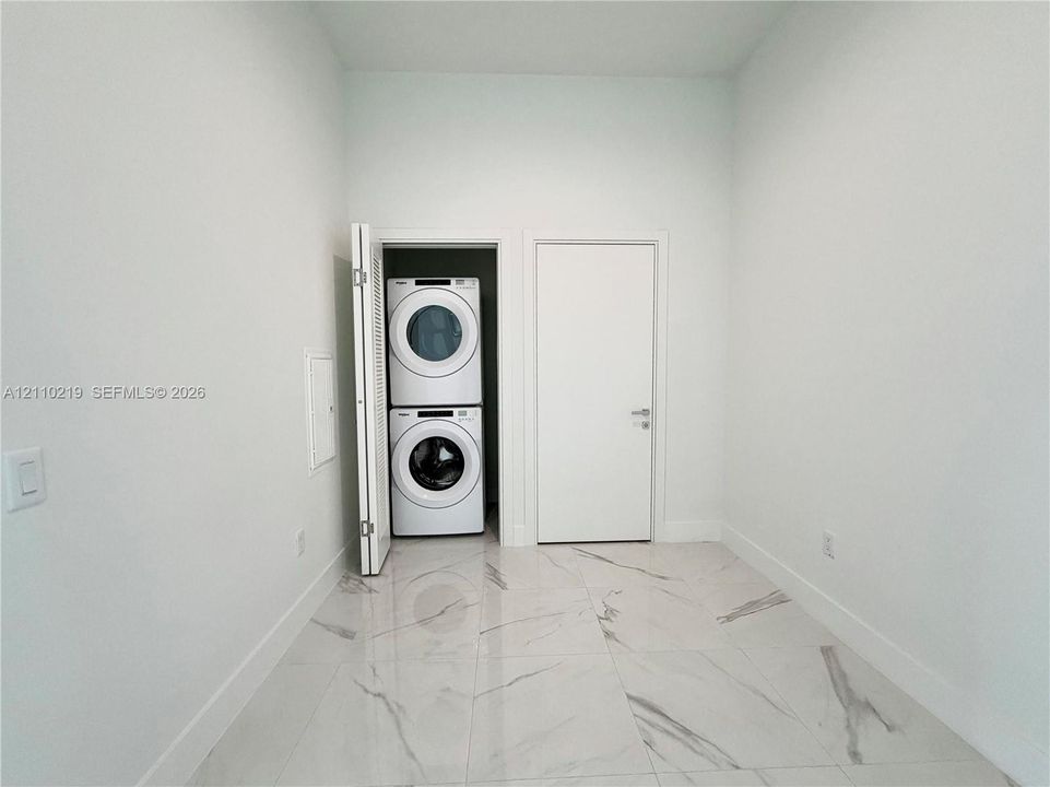 Utility area featuring large-format tile flooring with a neutral veined pattern, crisp white walls, and a recessed closet housing a stacked washer and dryer unit