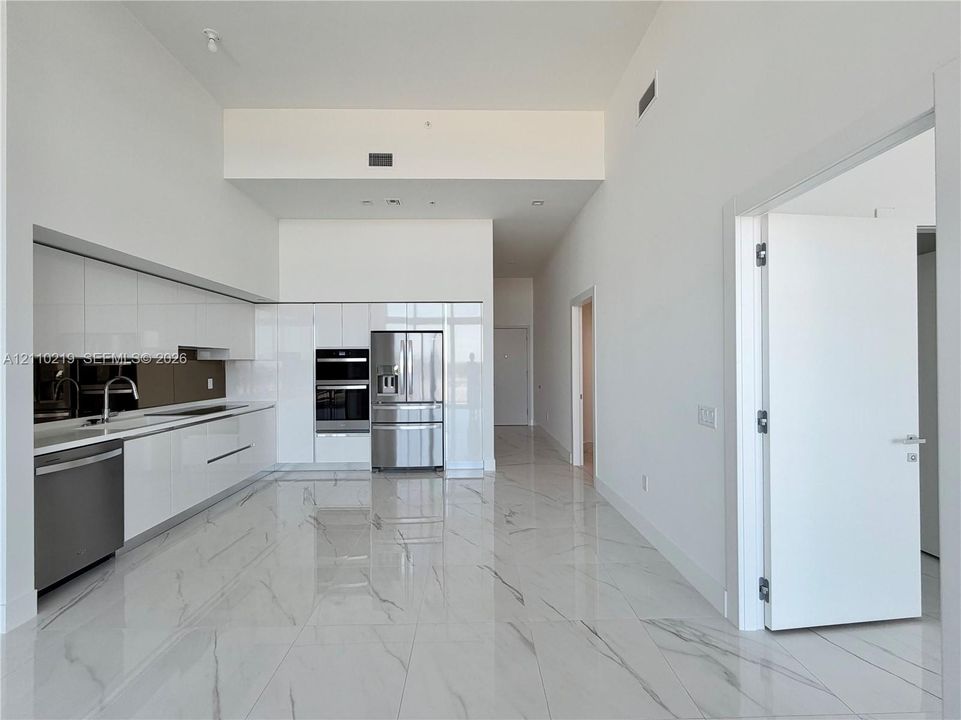 Contemporary kitchen featuring high-gloss white cabinetry, integrated stainless steel appliances, polished stone-finish tile flooring, and soaring ceilings