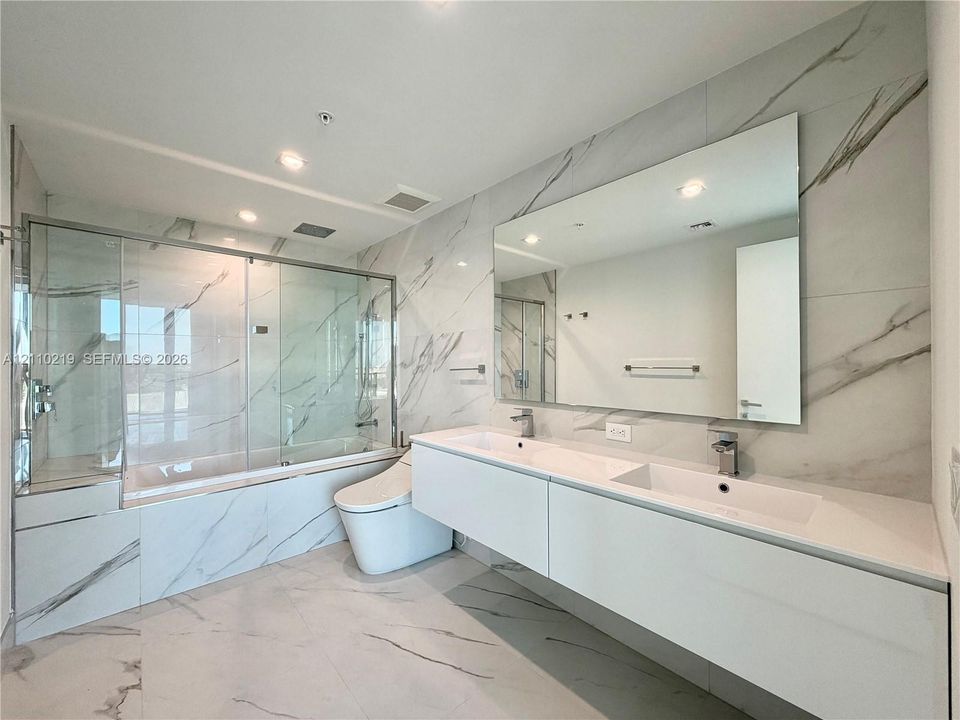 Floating dual-sink vanity with a solid surface countertop, expansive wall-to-wall mirror, glass-enclosed shower and tub combination, and large-format veined tile surround