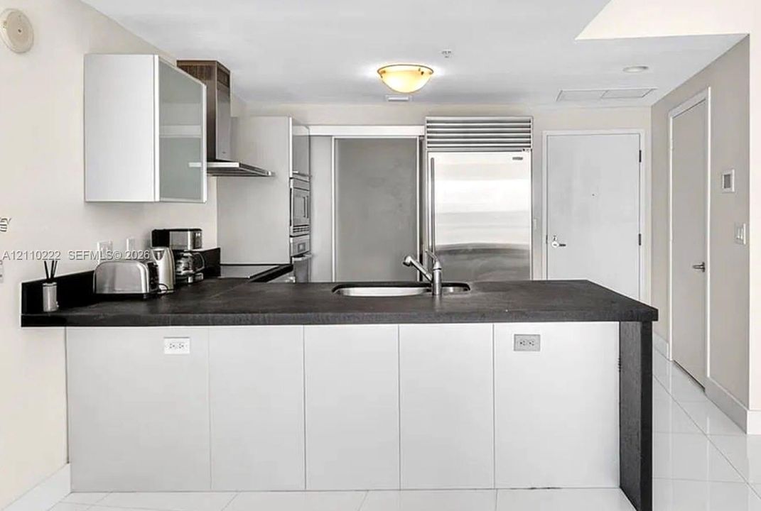 Kitchen featuring flat-panel cabinetry, a stainless steel refrigerator, stone-surfaced island with an integrated sink, and stainless steel range hood