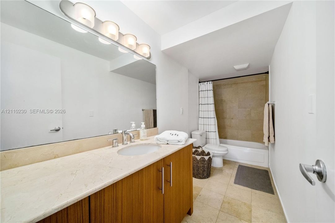 Bathroom featuring a vanity with a light-toned stone countertop, wood-finish cabinetry, a large wall-mounted mirror, four-bulb vanity lighting, and a bathtub with tile surround