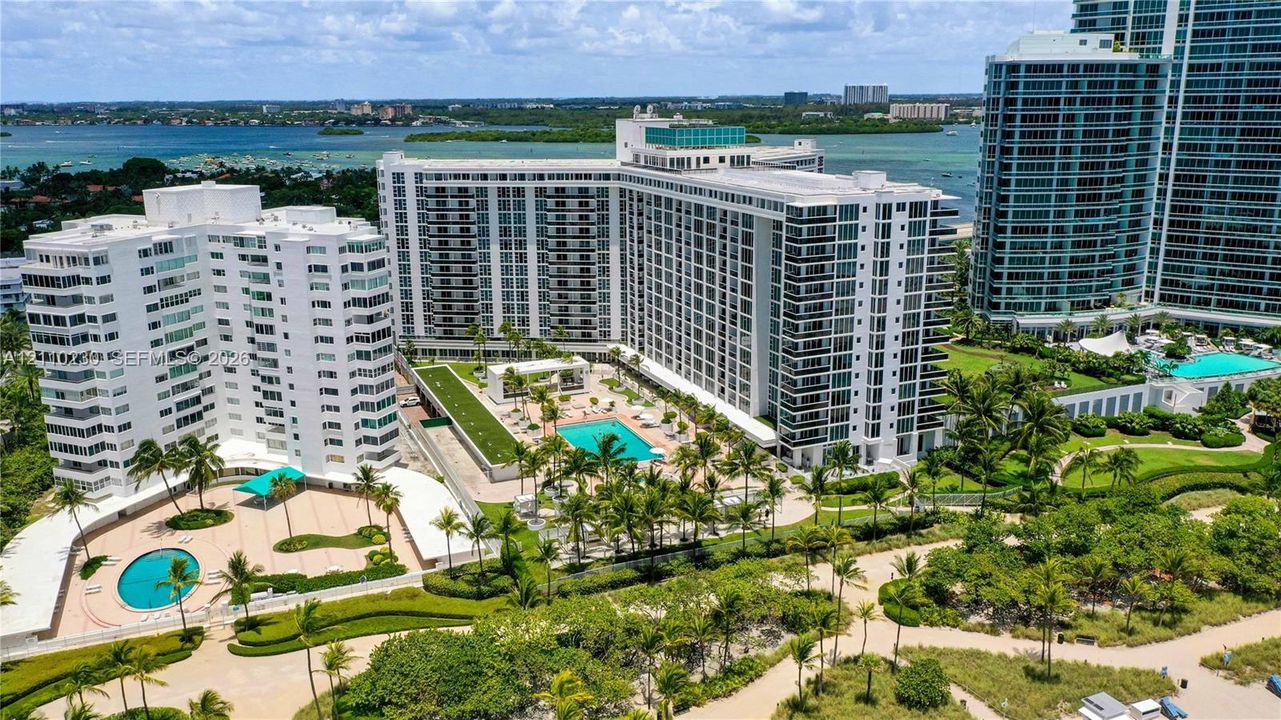 Aerial perspective of residential high-rise buildings featuring multiple swimming pools, expansive paved sun decks, manicured landscaping with palm trees, and proximity to coastal water