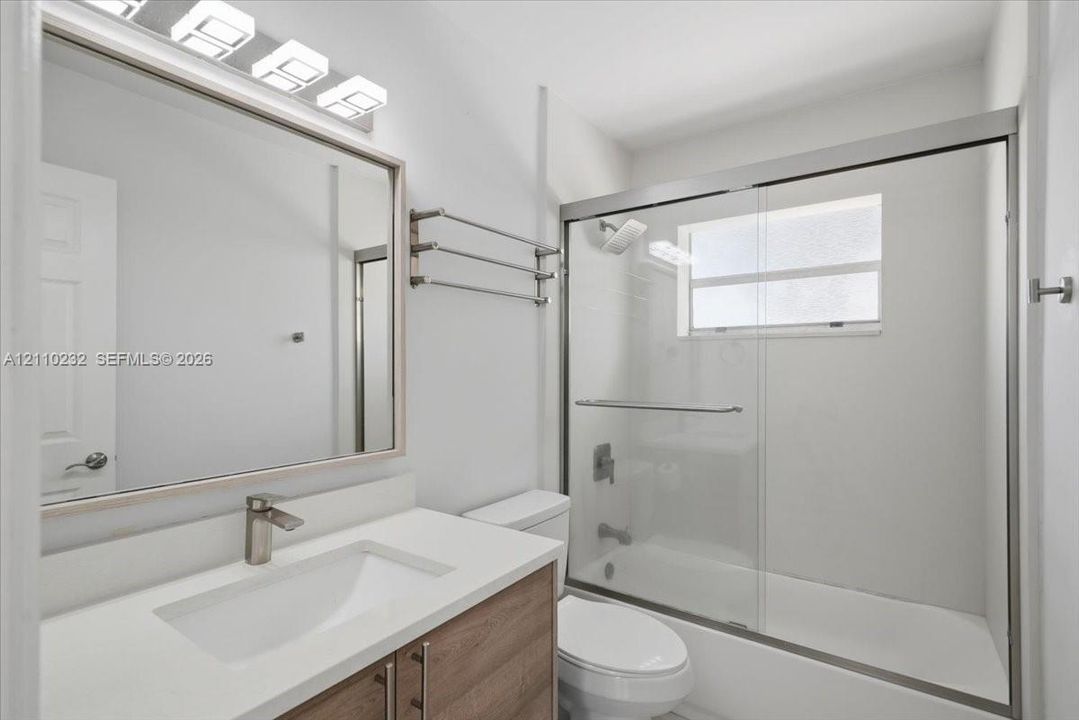 Wood-finish vanity with white countertop, integrated sink, brushed-metal hardware, sliding glass shower enclosure, and three-light wall-mounted fixture