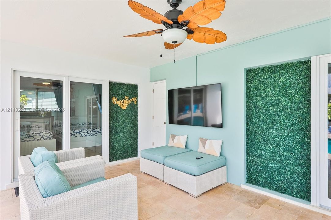 Covered outdoor living space featuring stone flooring, a ceiling fan with leaf-style blades, and decorative faux-greenery wall panels