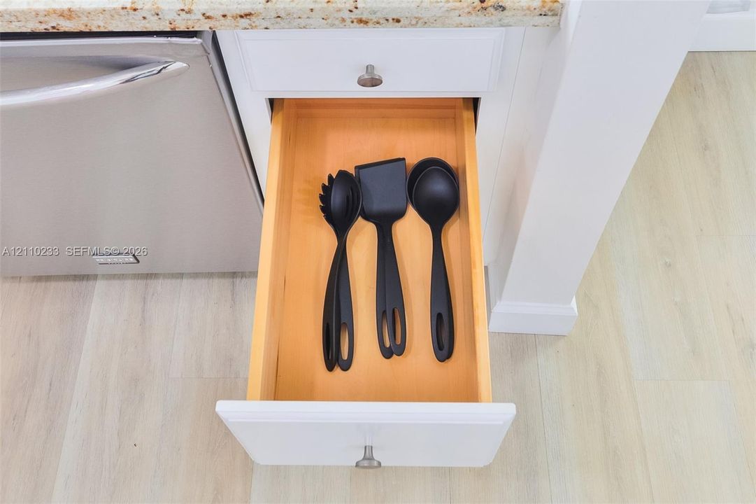 Custom-fit cabinetry featuring a pull-out drawer with a natural wood interior, white shaker-style paneling, a stainless steel dishwasher, and stone-surfaced countertops