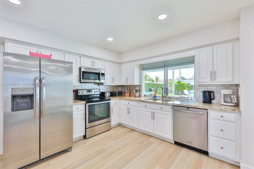 Kitchen featuring white shaker-style cabinetry, light-toned stone countertops, subway tile backsplash, wood-finish flooring, and stainless steel appliances