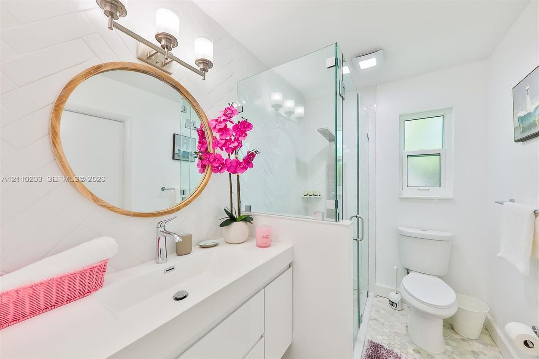 Bathroom featuring a wall-mounted vanity with integrated sink, glass-enclosed shower, round mirror with wood frame, three-light vanity fixture, and herringbone-patterned tile wall