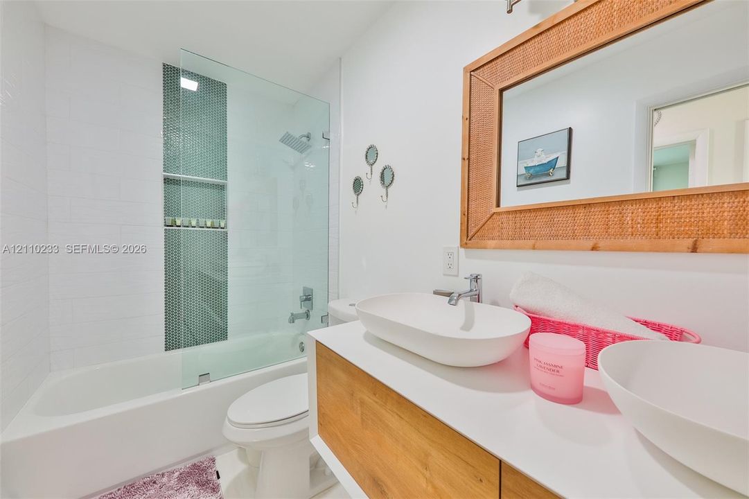 Bathroom featuring a wood-finish floating vanity with dual vessel sinks, a glass-enclosed tub shower with mosaic tile inlay, and a built-in wall mirror
