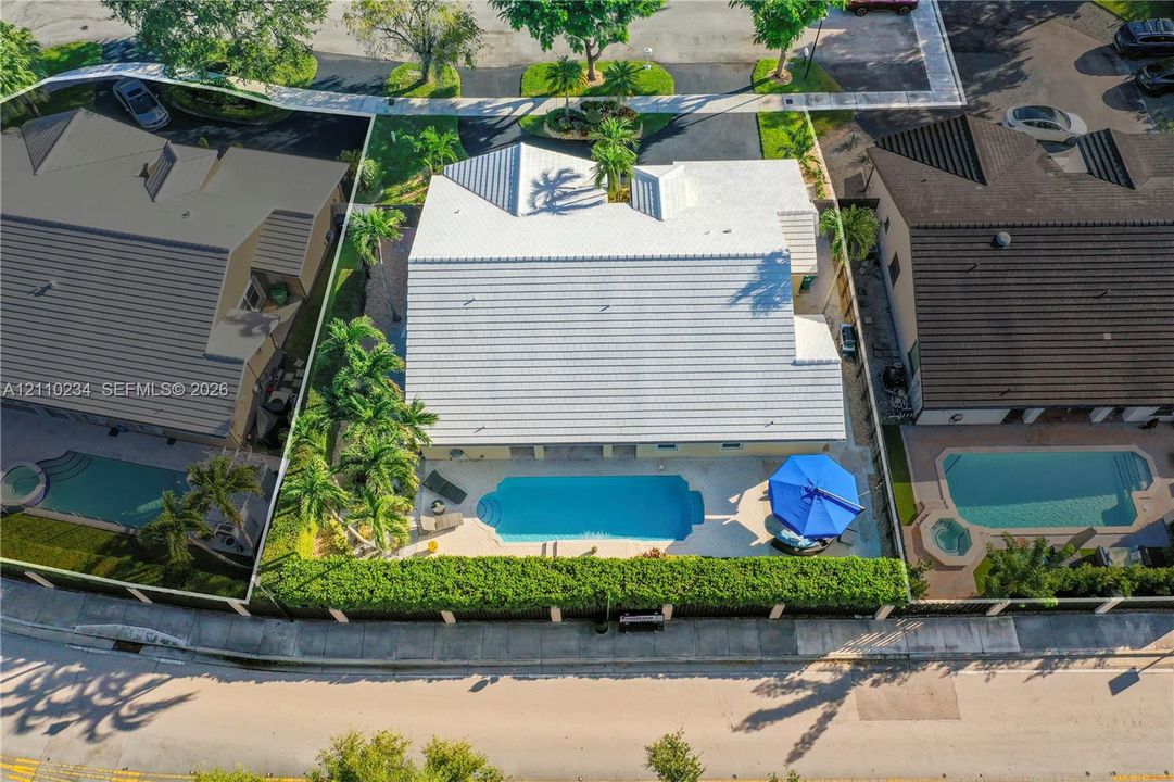 Aerial perspective of property featuring a light-toned tile roof, an in-ground swimming pool with a hardscaped deck, and mature palms within a fenced perimeter