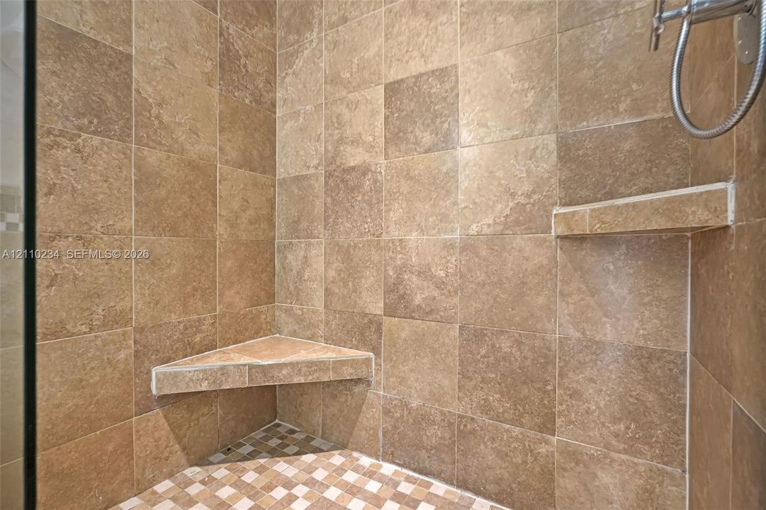 Walk-in shower featuring large-format neutral-toned tile walls, a built-in corner bench, a floating corner shelf, and a mosaic-tiled floor with metal shower fixture