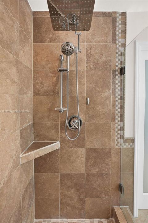 Walk-in shower featuring large-format earth-tone tile, a square rainfall showerhead with a secondary handheld sprayer, a built-in triangular corner shelf, and a vertical mosaic accent border