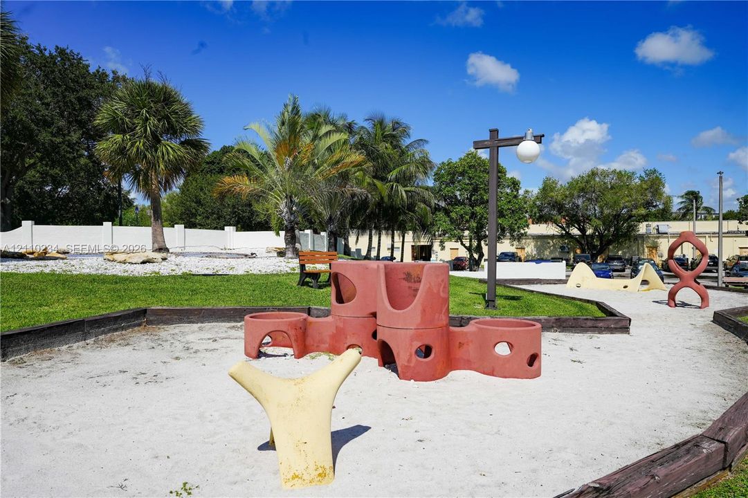 Recreational area featuring a sand-surfaced playground with sculpted climbing structures, manicured lawn, mature palm trees, and perimeter fencing
