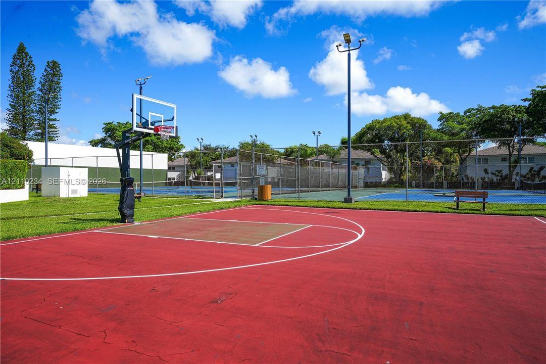 Outdoor recreation area featuring a paved basketball court, fenced tennis court, perimeter lighting, and surrounding green space