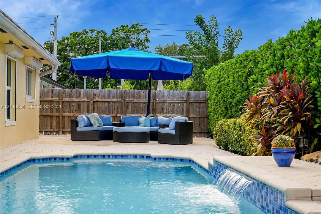 In-ground swimming pool featuring a decorative water feature and paver stone decking enclosed by a privacy fence and mature perimeter landscaping