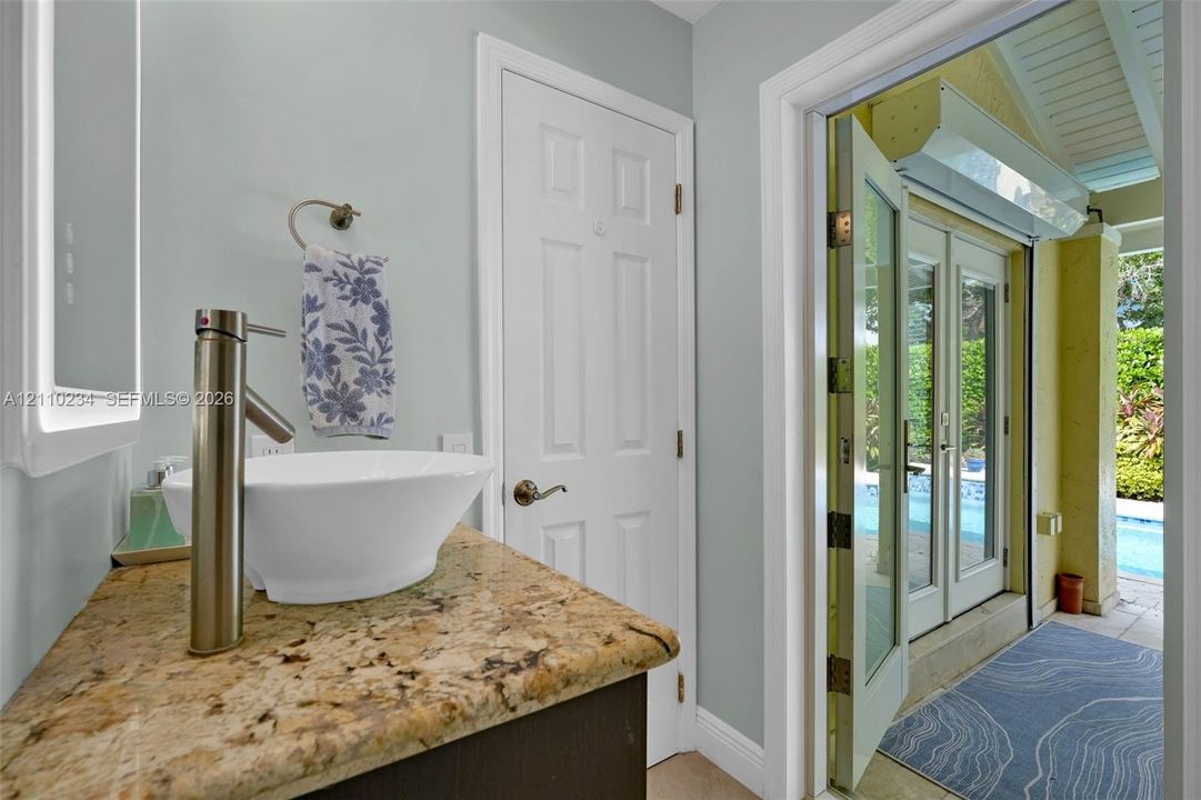 Vanity featuring a stone countertop, vessel sink with a high-arch faucet, and an integrated LED-backlit mirror, with double French doors opening to an exterior pool area