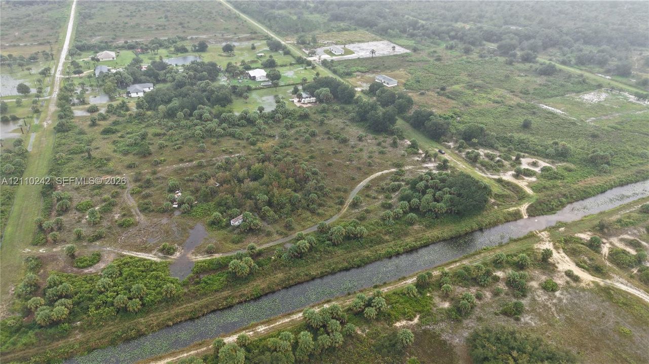 Aerial perspective of expansive acreage featuring native vegetation, clusters of mature trees, dirt pathways, and a canal